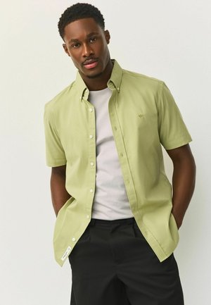 Vīrietis ar rokām kabatās valkā gaiši zaļu īsroku pogājamu kreklu virs balta T-krekla un melnas bikses, stāvot pret vienkāršu fonu.