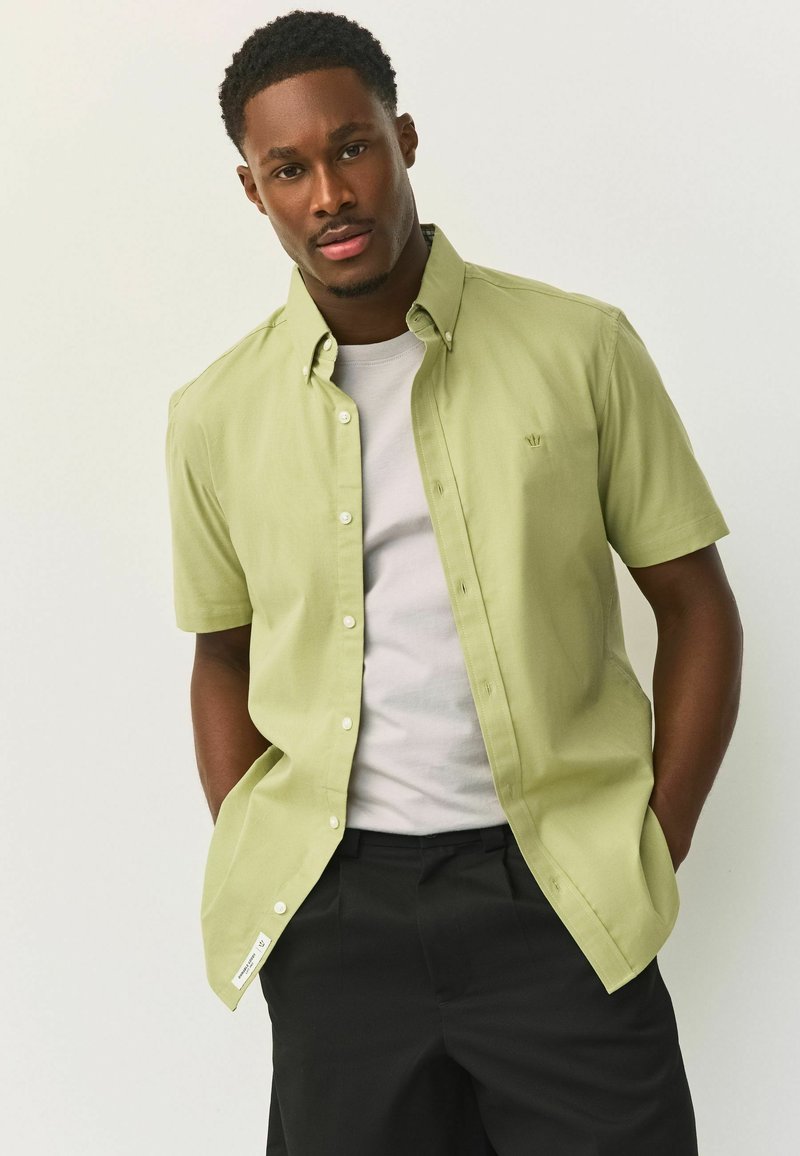 Mees, kellel on käed taskutes, kannab heledat rohelist lühikeste varrukatega nööbitavat särki valge t-särgi ja mustade pükste peal, seistes ühevärvilise tausta ees.