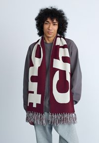 Cachecol bordô com texto branco em negrito, bordas franjas e uma textura suave, usado sobre um moletom cinza e calças cinzas claras.
