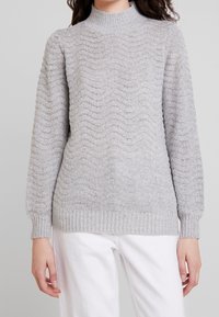Pull en maille gris clair avec un motif texturé en forme de vague, col montant, poignets et ourlet côtelés. Tissu doux de poids moyen.