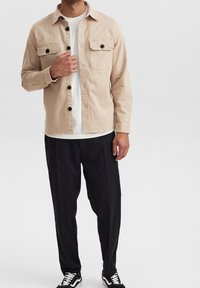 Vaalean beige nappikauluksinen takki, jossa on kaksi rintataskua, päällä valkoinen T-paita. Mustat löysät housut ja musta-valkoiset lenkkarit täydentävät asua.