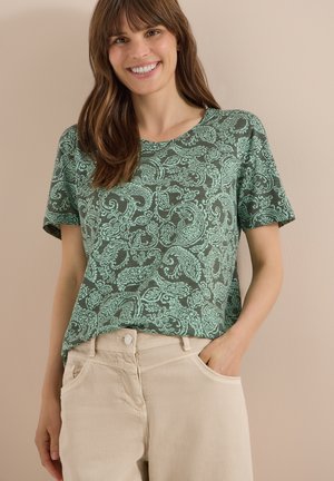 Femme souriante portant une chemise verte à manches courtes à motif cachemire et un pantalon beige, une main dans la poche, sur fond beige.