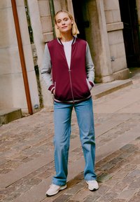 Jeune femme se tenant sur une rue en pavés, portant une veste zippée bordeaux et grise, un jean bleu et des baskets blanches, les mains dans les poches.