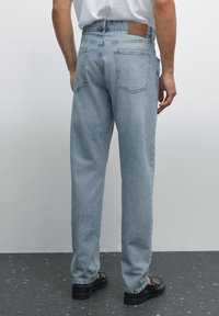 Light blue denim jeans with a relaxed fit, rear patch pocket detailing, and a leather brand tag on the waistband, worn with black shoes.