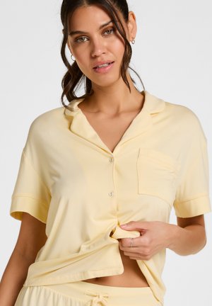 Frau mit dunklen Haaren, die ein blassgelbes kurzärmeliges Hemd und passende Hose trägt, hält mit einer Hand den Saum des Hemdes.