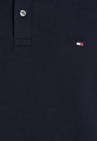 Dunkel marineblaues Poloshirt mit silbernem Knopf und kleinem besticktem rot-weiß-blauem Flaggenlogo auf der Brust.