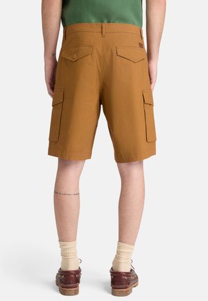 Mężczyzna ubrany w brązowe szorty bojowe, beżowe skarpetki i brązowe buty, stoi tyłem na białym tle.