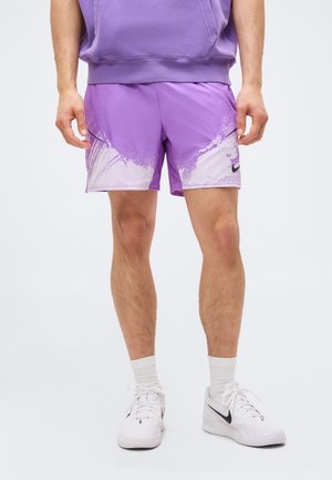 Person, der bærer lilla og hvide sportsshorts med splashmønster, hvide sportssko, hvide sokker og en lilla hættetrøje.