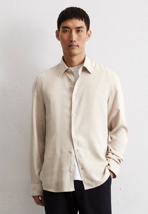 Beige overhemdd met een kraag, lange mouwen en een gladde textuur. Model draagt het over een wit shirt en zwarte broek.