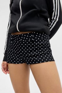 Frau in schwarzer Zip-Jacke mit weißen Streifen an den Ärmeln und schwarzen Shorts mit weißen Punkten, die vor einem schlichten Hintergrund steht.