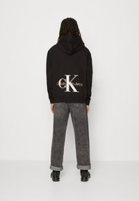 Sudadera con capucha negra que presenta un gran logo en blanco y oro "CK" en la parte posterior, combinada con unos vaqueros grises holgados y zapatos negros.