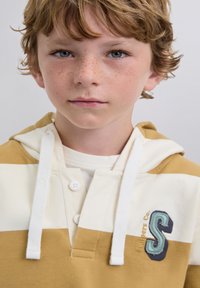 Niño pequeño con cabello castaño claro y pecas, que lleva una sudadera a rayas amarilla y blanca con un gran parche en forma de "S" en el pecho.