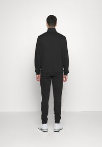 Black athletic tracksuit with a zip-up jacket and tapered pants, featuring a smooth texture and minimal design. Paired with white sneakers.