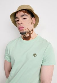 Tricou din bumbac de culoare verde mentă, cu mâneci scurte, având un mic logo maro în formă de copac pe partea stângă a pieptului; purtat cu o pălărie de tip bucket de culoare bej.