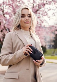 Black leather wallet with a rounded shape and snap closure, held in a hand, set against a backdrop of pink flowering trees.