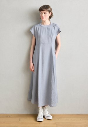 Helles graues, knöchellanges Kleid mit kurzen Ärmeln, gerafftem Bund und Taschen. Getragen mit weißen High-Top-Sneakern auf einem Holzboden.