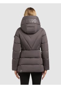 Naine kannab halli paksu talvejopet, millel on suur kapuuts, millel on "KHUJO" tekst, vaadatuna seljaga ühtlase tausta ees.