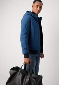 Blue, hooded jacket with a zip front and ribbed cuffs, paired with denim jeans. Holding a large, textured black duffle bag.