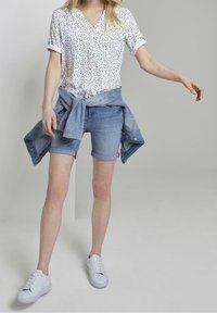 Weiße Kurzarmbluse mit schwarzen Polka-Dots, kombiniert mit hellen Denim-Shorts und blauem Denim, der an der Taille gebunden ist. Weiße Turnschuhe vervollständigen den Look.