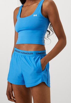 Kvinde iført blå Under Armour ærmeløs crop top og matchende atletiske shorts, med den ene hånd i lommen mod en ensfarvet baggrund.