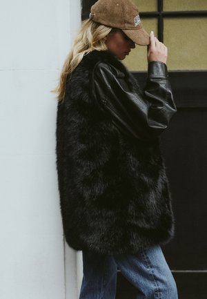 Svart faux fur ärmlös kappa över en svart lädertröja med långa ärmar, tillsammans med blå denimjeans och en brun manchesterkeps.