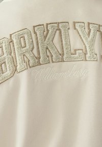 Tessuto beige con lettere "BRKLYN" ricamate in crema e la scritta corsiva "Williamsburg" sottostante. Texture morbida e liscia.