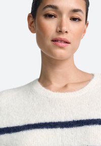 Fehér kötött pulóver puha textúrával, sötétkék vízszintes csíkkal a mellkason és kerek nyakkivágással.