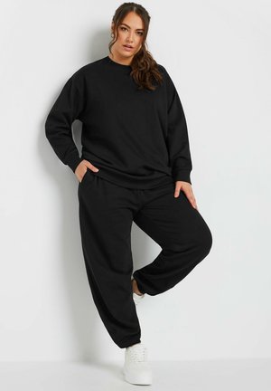 Kvinde iført sort sweatshirt og joggers, stående med den ene hånd i lommen og det ene ben bøjet, foran en ensfarvet hvid baggrund.