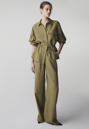 Kobieta ubrana w oliwkowozieloną, długą koszulę zapinaną na guziki z taliowanym sznurkiem oraz dopasowane szerokie spodnie, stoi z jedną ręką w kieszeni.