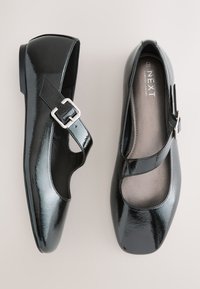 Paar schwarze Mary Jane Lacklederschuhe mit silbernen Schnallen und gepolsterten Innensohlen, von oben auf hellem Hintergrund gezeigt.