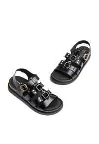 Sandalias de cuero negro con múltiples tiras anchas, hebillas doradas y acentos de tachuelas. Suela plana con un plantillaje texturizado de corcho. Diseño de apertura en la parte trasera.