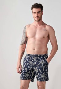 Marineblå svømmebukser med et hvidt blomstermønster, elastisk talje og sidelommer. Stoffet har en glat tekstur og en afslappet pasform.