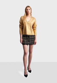 Guldig metallisk långärmad topp med V-ringning, kombinerad med en svart och guldig mönstrad minikjol med knappar och fransade kanter. Svarta klackar.