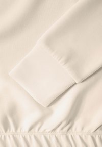 Tessuto beige chiaro con una trama liscia. Presenta una manica con polsini e un orlo arricciato con dettagli di cucitura visibili, che enfatizzano l'elasticità.