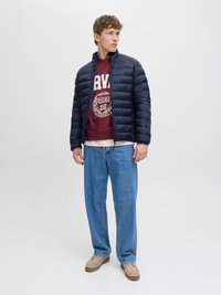 Jūrų laivyno pūkinė striukė su stovinčia apykakle, raudono vyno spalvos džemperis su baltu tekstu, laisvi šviesiai mėlyni džinsai ir smėlio spalvos šliaužikliai.