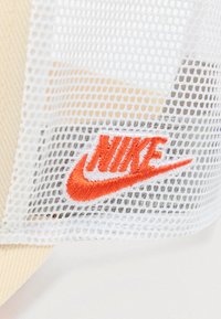 Bílá síťovaná tkanina s vyšitým oranžovým logem Nike a symbolem swoosh, částečně překrývající béžový texturovaný materiál.