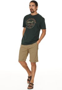 Dunkelgrünes Baumwoll-T-Shirt mit goldfarbener Logobeschriftung, kombiniert mit beigen Baumwollshorts. Gestylt mit braunen Sandalen, die ein Zwei-Riemen-Design aufweisen.
