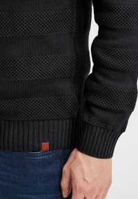 Maglione nero in maglia con strisce testurizzate distintive, polsini e orlo a costine, e una piccola etichetta in pelle marrone sul fondo.