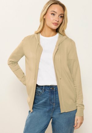 Femme portant un sweat à capuche beige zippé sur un t-shirt blanc et un jean bleu, posant avec une main sur la hanche devant un fond uni.
