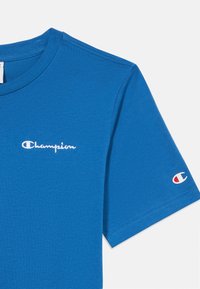 Μπλε βαμβακερό T-shirt με στρογγυλή λαιμόκοψη και κοντά μανίκια. Στο μπροστινό μέρος υπάρχει λευκό κεντημένο λογότυπο "Champion"· το αριστερό μανίκι έχει μια μικρή λογότυπο λεπτομέρεια.