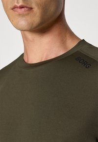 Közelkép egy személyről, aki olívazöld kereknyakú pólót visel, amin a "BORG" felirat látható a váll közelében.