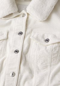 Vitt kordjacka med en mjuk, ljus krage. Har silverfärgade tryckknappar och två framfickor. Texturerat tygmönster.