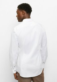 Weißes Langarmhemd mit spitzem Kragen, verziert mit einem geschwungenen Saum und geknöpften Manschetten, gefertigt aus glattem Stoff mit leichtem Glanz.