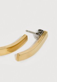 Auksaspalvės, išlenktos metalinės auskarai, turintys blizgų paviršių, stačiakampius kraštus ir išpjovą smeigtuko gale. Matoma išgraviruota prekės ženklas.