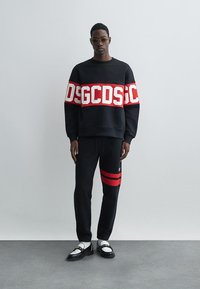 Μαύρη μπλούζα με έντονη κόκκινη επιγραφή "DSGCDS", συνδυασμένη με μαύρο παντελόνι που έχει κόκκινες ρίγες και μαύρα παπούτσια με λευκές λεπτομέρειες.