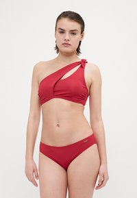 Rød bikini med ensidig design og en knyttet detalj, glatt tekstur og matchende lavt skårne bukser.