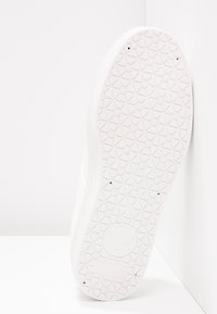 Semelle de sneaker en caoutchouc blanc présentant un motif géométrique texturé, avec un logo en relief au niveau du talon. Bords lisses avec un design minimaliste.