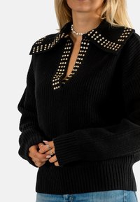 Maglione nero lavorato a maglia con orlo a costine spesso, colletto decorato con borchie d'oro e d'argento e scollo a V. Maniche lunghe e morbide.