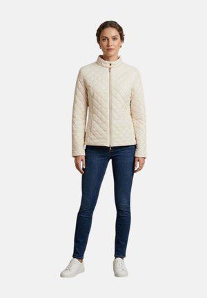 Donna con giacca trapuntata beige, jeans skinny blu scuro e sneakers bianche in piedi davanti a uno sfondo semplice.