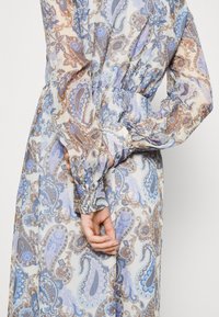 Pitkä, läpikuultava mekko sinisen ja ruskean sävyisen paisley-kuviolla. Mekossa on frillattu vyötärö ja puffihihat, joissa on napitettu hihansuu.